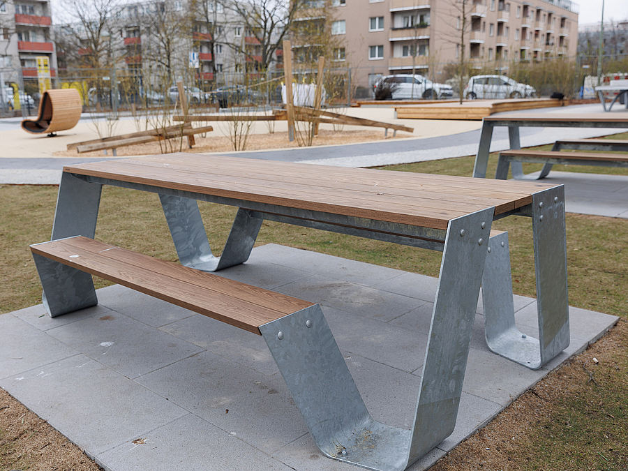 2 fest installierte Tisch-Bank-Kombinationen aus Metall und Holz, dahinter Sandspielplatz im Winter