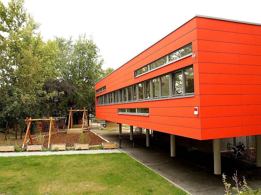 Rotes, aufgeständertes Gebäude mit Rasen, im Hintergrund Seilbahn