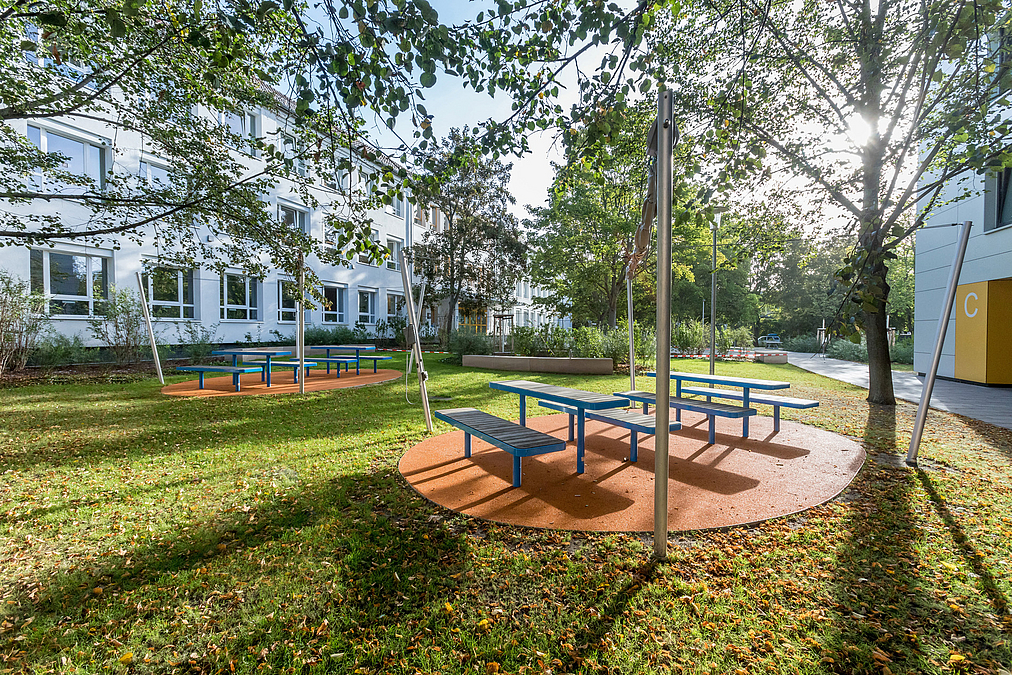 Ovale Kunststofffläche mit Tischen und Bänken unter Laubbäumen im Rasen zwischen 2 Gebäuden