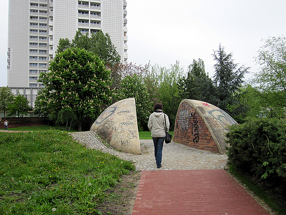 Frau geht im Park durch ein Kunstwerk in Form zweier Viertelkugeln, im Hintergrund Hochhaus