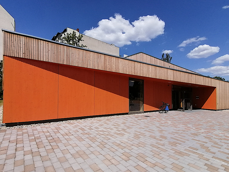 Eingeschossige, orangefarbene Front mit Eingang und Bodenfenster, darüber Holzlamellen, davor Pflaster