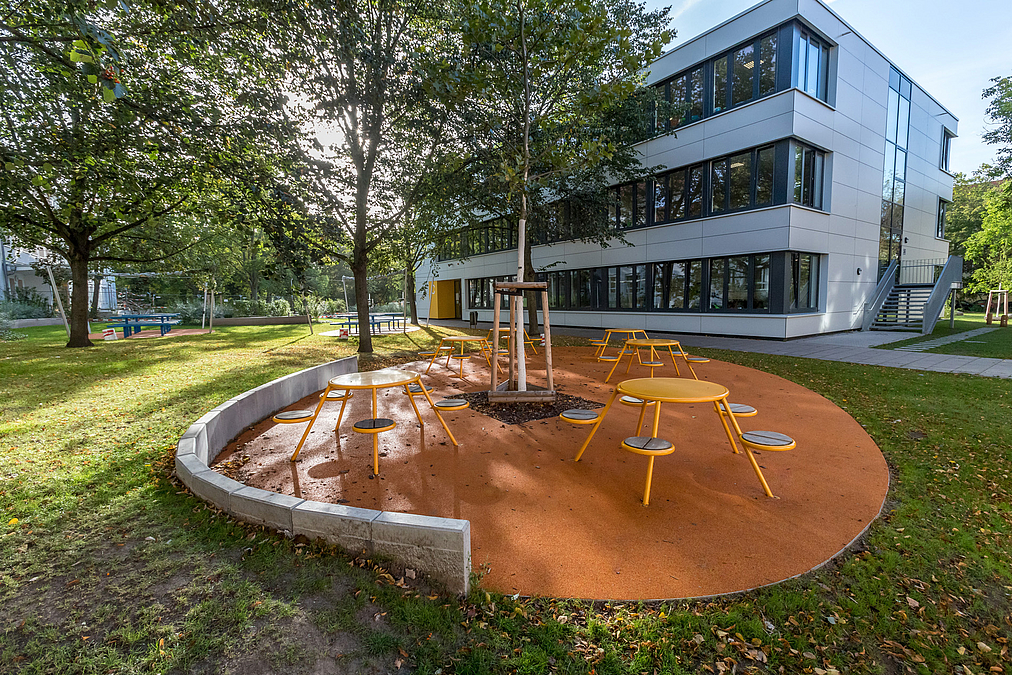 Helles Schulgebäude, Kunststofffläche mit mittigem Baum und festen Sitzgruppen mit runden Tischen und Hockern