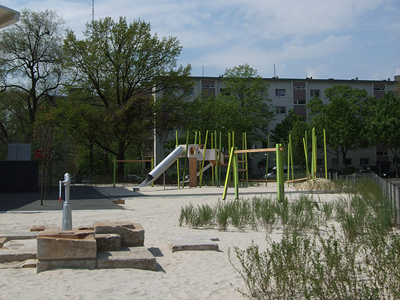 Sandspielplatz mit Pumpe zwischen Steinquadern, großem Klettergerät mit Rutsche und Büschen im Sand